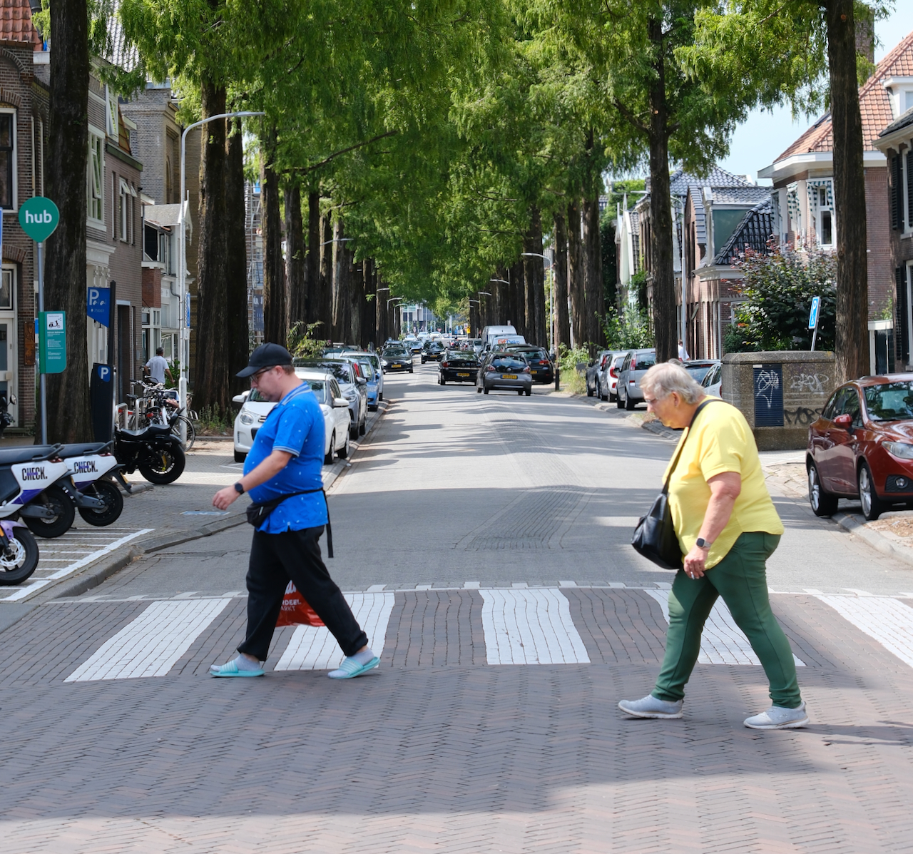 Two people crossing a tree-lined street on a bright summer day